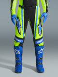 Alpinestars 2026 Techstar Nomur Pants, spodnie motocrossowe, żółte neonowe (fluo) i ciemnogranatowe z niebieskimi akcentami, noszone z dopasowanymi niebieskimi rękawicami i butami, wyraźne logo Astars na udach, wyczynowa konstrukcja do wyścigów terenowych
