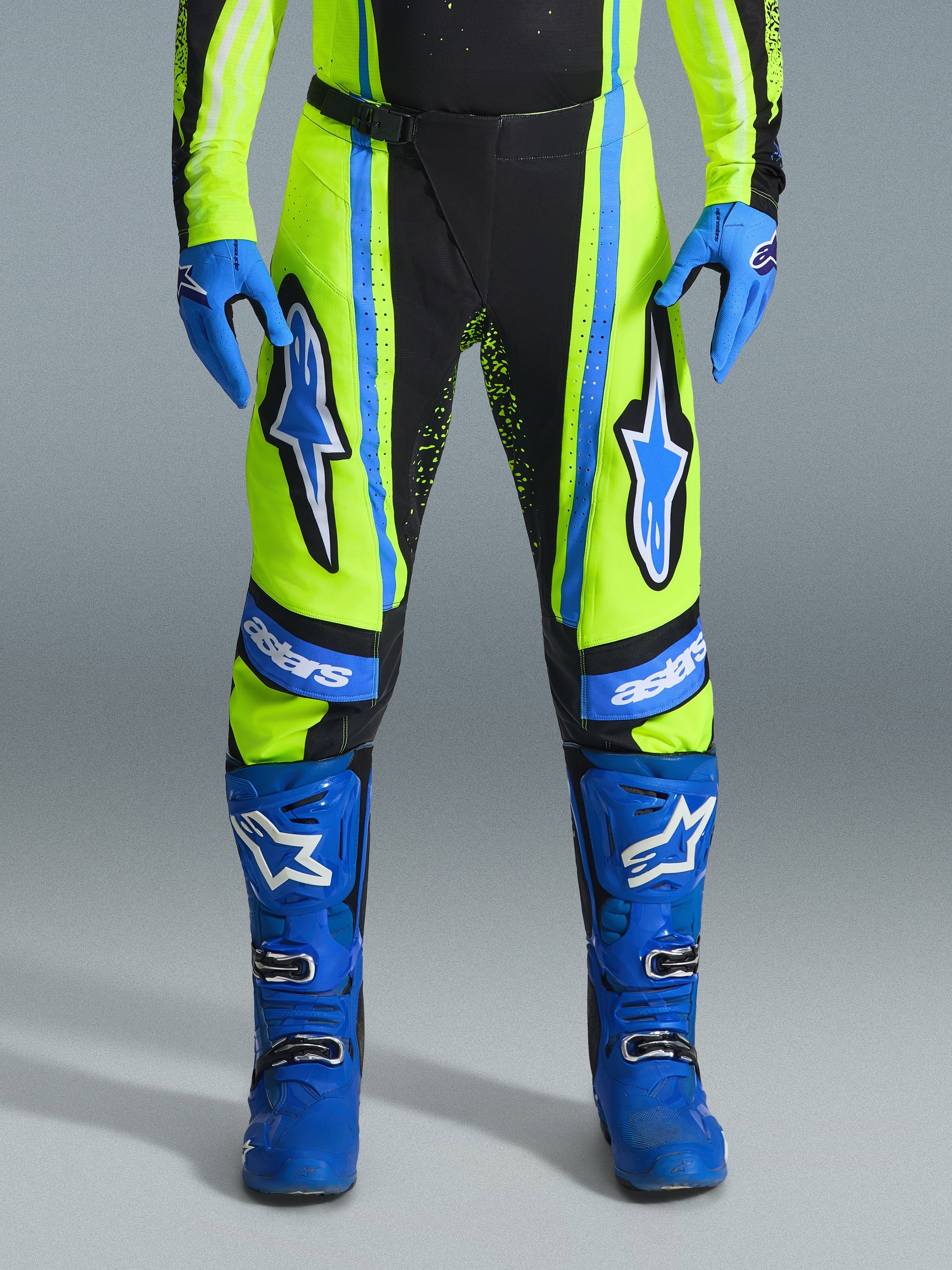 Alpinestars 2026 Techstar Nomur Pants, spodnie motocrossowe, żółte neonowe (fluo) i ciemnogranatowe z niebieskimi akcentami, noszone z dopasowanymi niebieskimi rękawicami i butami, wyraźne logo Astars na udach, wyczynowa konstrukcja do wyścigów terenowych
