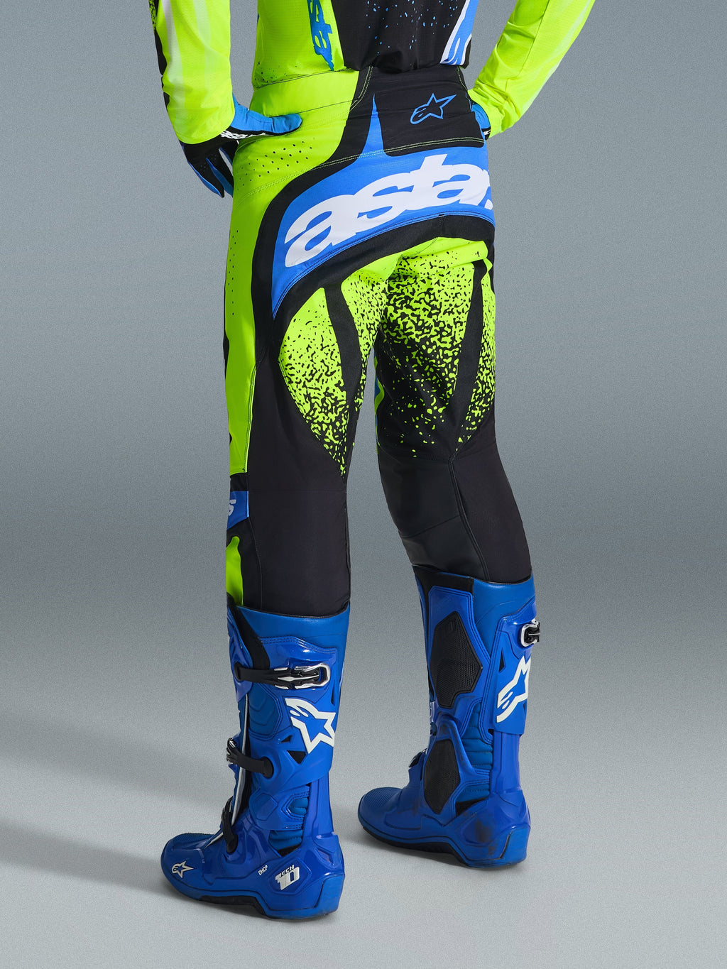 Alpinestars Techstar Nomur Pants 2026, spodnie motocrossowe, żółty neonowy i niebieski UCLA Blue, widok z tyłu na motocyklistę w niebieskich butach, z brandingiem „astars” na siedzisku i czarnymi grafikami z motywem odprysków na nogawkach