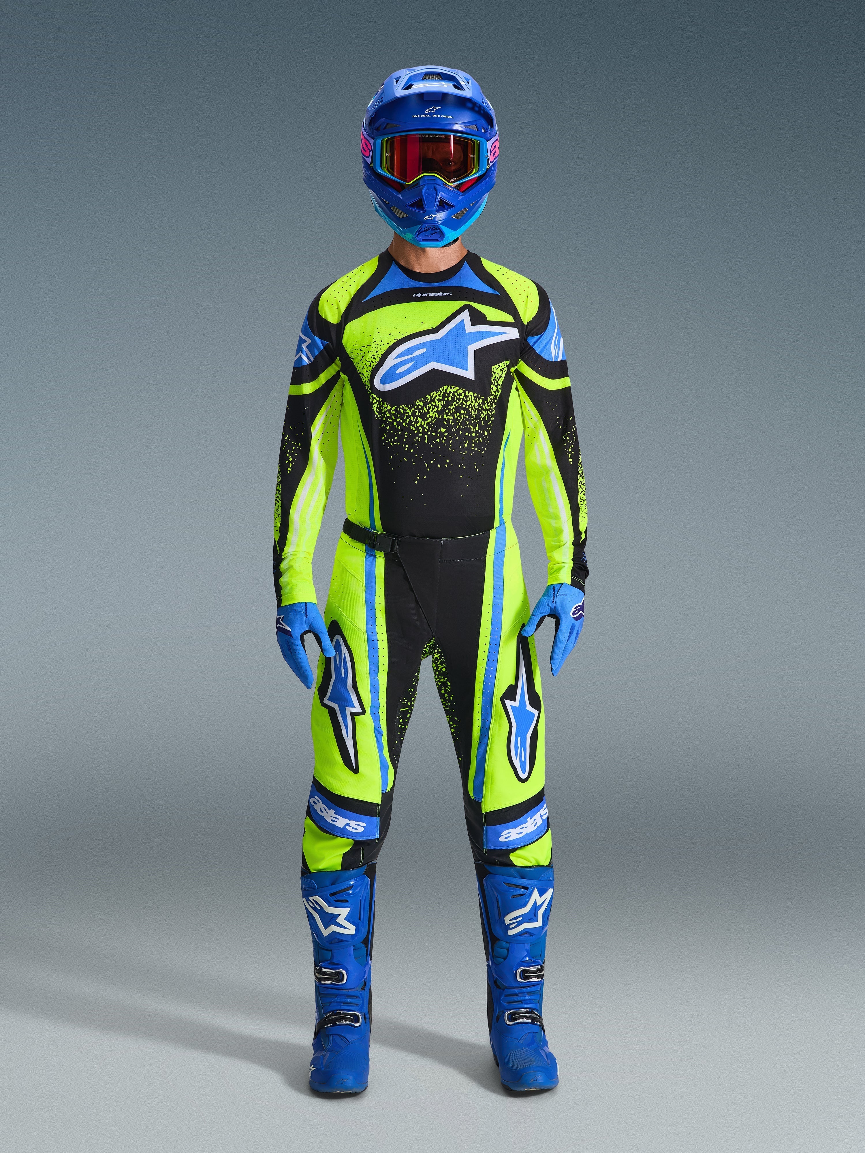 Zawodnik motocrossowy w pełnym stroju wyścigowym, spodnie i bluza Alpinestars 2026 Techstar Nomur w kolorach żółtym fluo i ciemnogranatowym z niebieskimi akcentami i logo, w zestawie z niebieskim kaskiem, rękawicami i butami dla spójnego wyglądu wyścigowego off-road.
