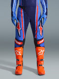 Alpinestars 2026 Techstar Nomur Pants, spodnie motocrossowe, ciemnogranatowe, pomarańczowe i błękitne UCLA, z wyrazistymi pomarańczowymi pasami i dużym białym logo gwiazdy na udach, noszone z jaskrawopomarańczowymi butami wyścigowymi i niebieskimi rękawicami, sportowy krój wyścigowy