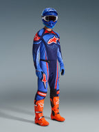 Alpinestars 2026 Techstar Nomur Pants, spodnie motocrossowe, ciemnogranatowe, pomarańczowe i błękitne (UCLA Blue), noszone przez zawodnika w pełnym stroju wyścigowym, w tym pasująca koszulka, niebieski kask i jaskrawopomarańczowe buty, zaprojektowane do profesjonalnych wyścigów terenowych