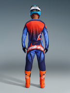 Motocyklista motocrossowy widziany od tyłu w spodniach Alpinestars 2026 Techstar Nomur w kolorach Dark Navy, pomarańczowym i UCLA Blue, zestawione z pasującą koszulką i jaskrawopomarańczowymi butami, profesjonalny zestaw odzieży do wyścigów terenowych