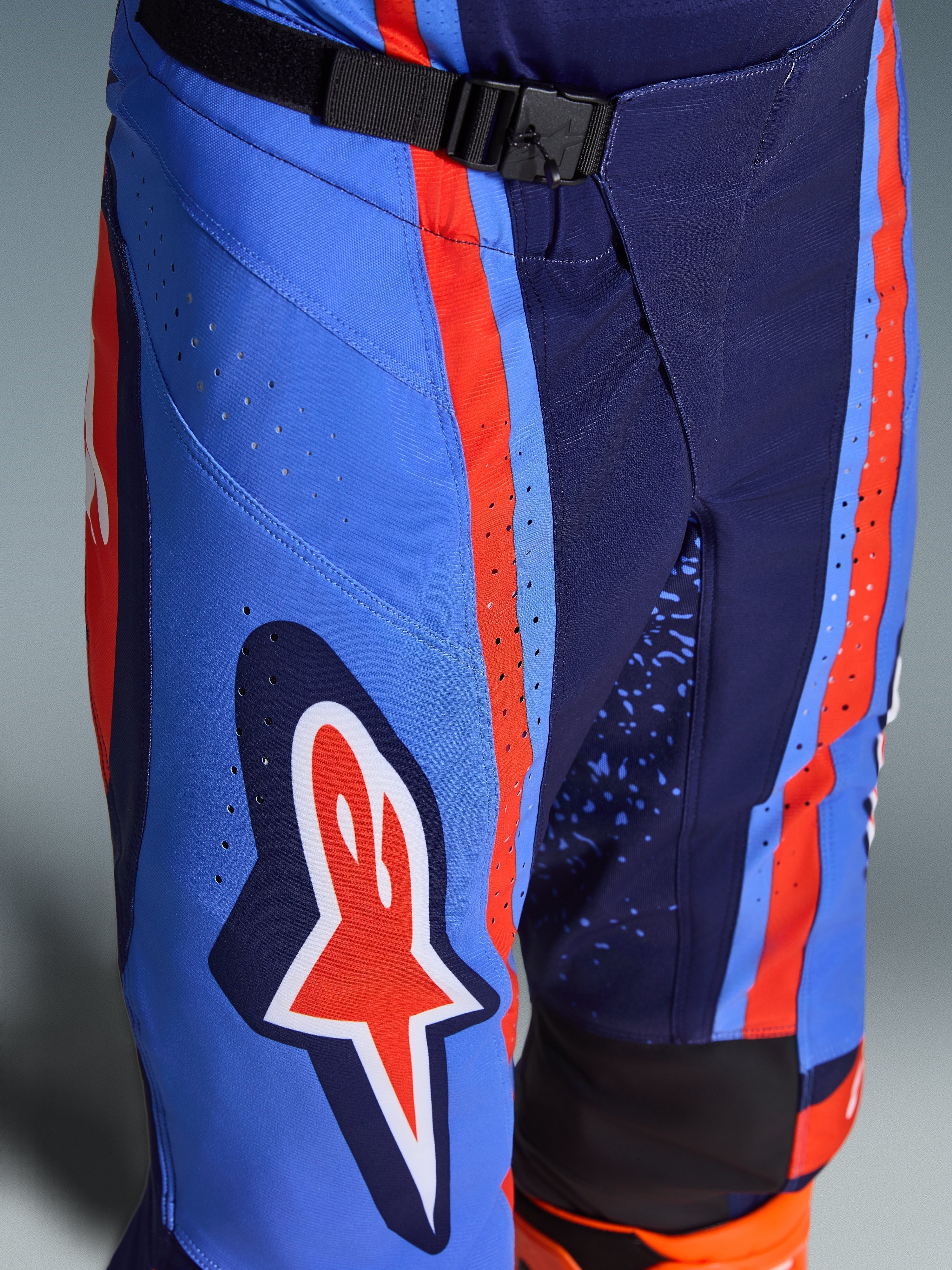 Alpinestars Techstar Nomur Pants 2026, spodnie motocrossowe w kolorach ciemnogranatowym, pomarańczowym i błękitnym, zbliżenie na magnetyczne zapięcie SX2 w pasie, laserowo wycinane perforacje na udzie oraz kultowe logo gwiazdy, wytrzymała odzież off-road