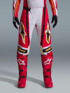Alpinestars Techstar Nomur Pants 2026, spodnie motocrossowe, czerwono-białe z czarnymi i żółtymi akcentami, noszone z pasującymi czerwonymi butami i czarnymi rękawicami, wyczynowa odzież wyścigowa off-road