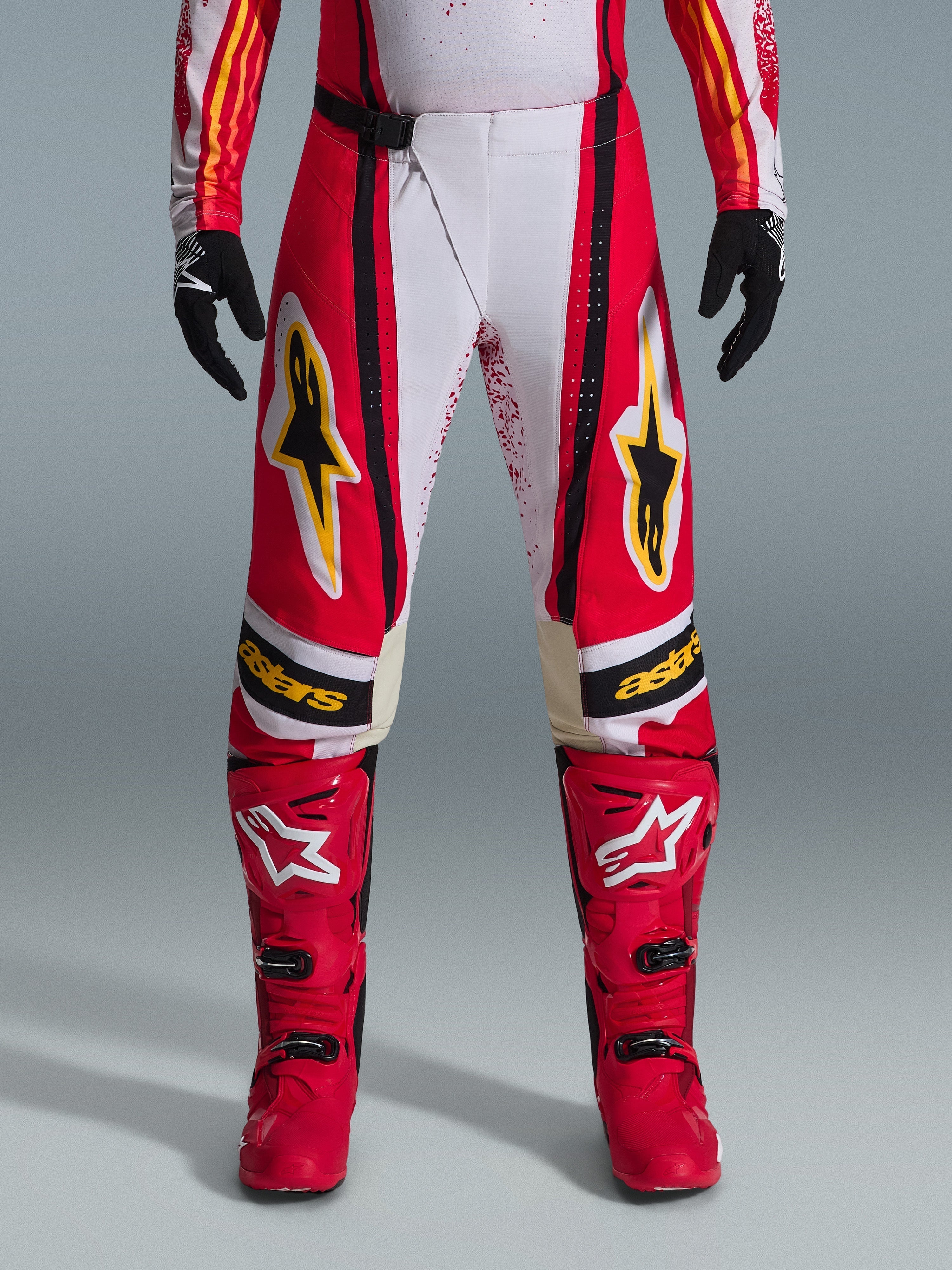 Alpinestars Techstar Nomur Pants 2026, spodnie motocrossowe, czerwono-białe z czarnymi i żółtymi akcentami, noszone z pasującymi czerwonymi butami i czarnymi rękawicami, wyczynowa odzież wyścigowa off-road