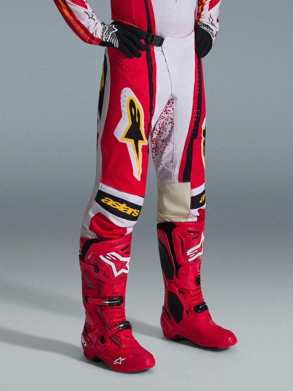 Alpinestars 2026 Techstar Nomur Pants, spodnie motocrossowe, czerwono-biało-czarne z żółtymi akcentami, noszone z dopasowanymi czerwonymi butami motocrossowymi i czarnymi rękawicami, żółte logo Alpinestars i napis „astars” na kolanach, profesjonalny sprzęt off-road.