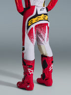Widok od tyłu motocyklisty motocrossowego, spodnie Alpinestars 2026 Techstar Nomur, czerwono-białe z wyraźnym żółtym logo na siedzeniu, jaskrawoczerwone buty motocrossowe Tech 10, zdjęcie studyjne na szarym tle.