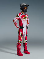 Alpinestars Techstar Nomur Pants 2026, spodnie motocrossowe, czerwono-białe z czarnymi i żółtymi akcentami, noszone przez motocyklistę z pasującą koszulką wyścigową, jaskrawoczerwonymi butami i czarnym kaskiem ze złotymi goglami, kompletny profesjonalny strój na zawody off-road