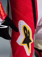 Alpinestars 2026 Techstar Nomur Pants, spodnie motocrossowe, czerwone z białymi i żółtymi akcentami, zbliżenie na obszar uda z laserowo wycinanymi otworami wentylacyjnymi i dużym logo gwiazdy, w zestawieniu z czarnymi rękawicami i kolorową koszulką off-road
