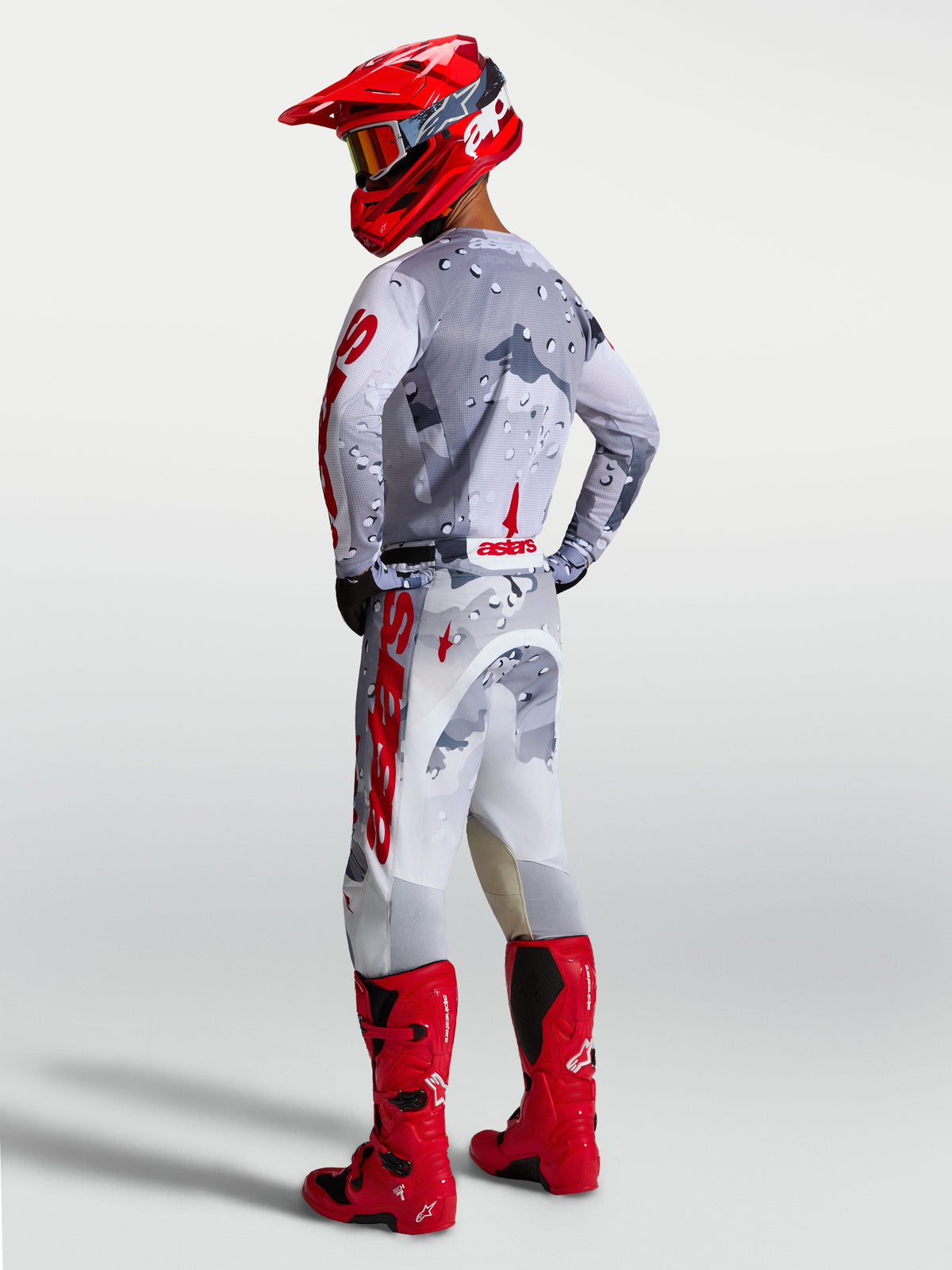 Alpinestars Racer Air Lone, męskie spodnie motocrossowe, szary kamuflaż i czerwony, wentylowana konstrukcja off-road z elastycznymi wstawkami, widok z tyłu na motocyklistę w pełnym ekwipunku.