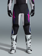 Alpinestars 2026 Fluid Grid Pants, spodnie motocrossowe, czarne z fioletowymi i niebieskimi wzorami gwiazd, noszone z białymi butami motocrossowymi i czarnymi rękawicami, wzmocnione panele, sportowy krój off-road, szare tło
