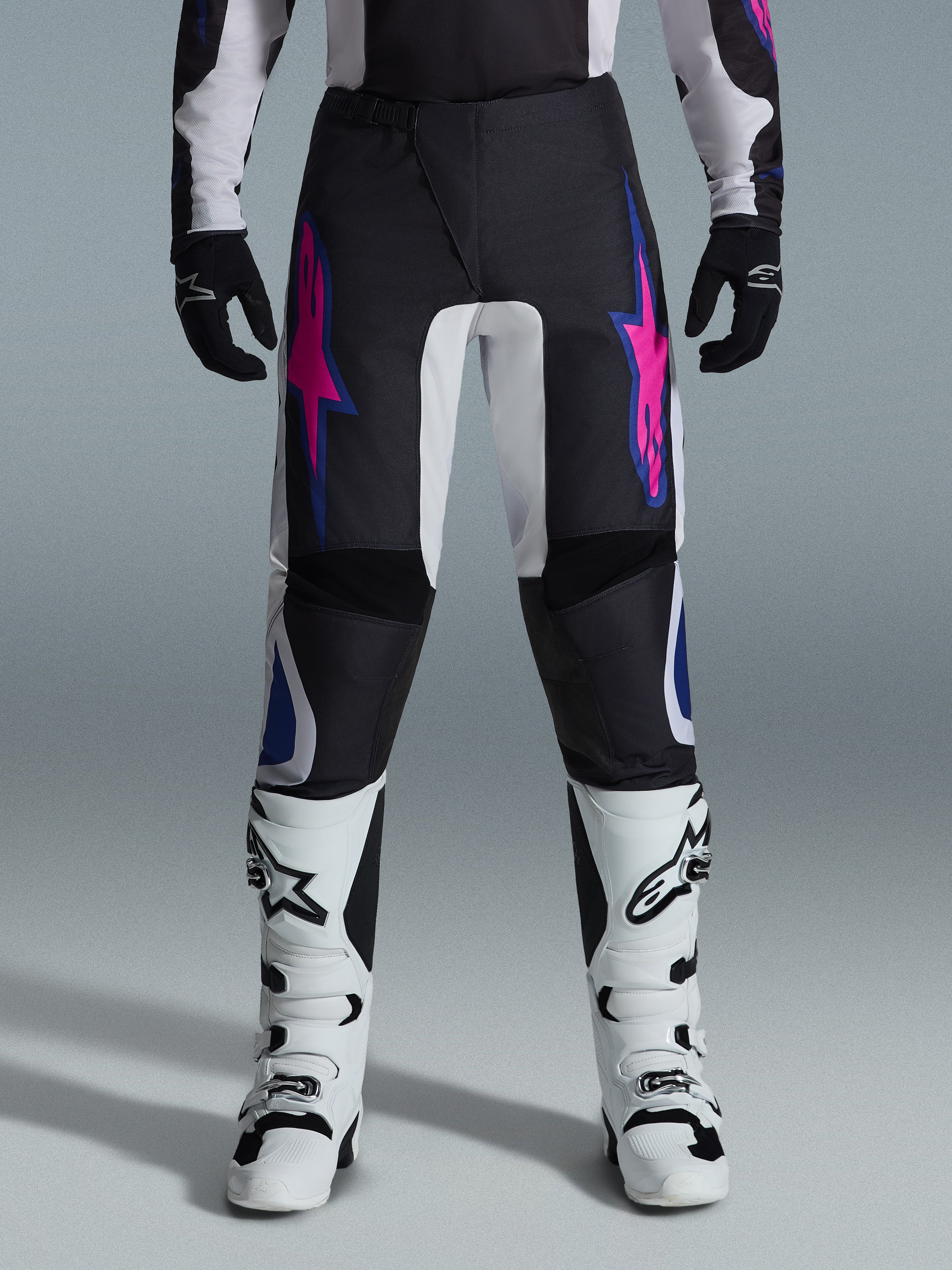 Alpinestars 2026 Fluid Grid Pants, spodnie motocrossowe, czarne z fioletowymi i niebieskimi wzorami gwiazd, noszone z białymi butami motocrossowymi i czarnymi rękawicami, wzmocnione panele, sportowy krój off-road, szare tło