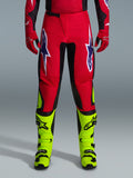 Spodnie Alpinestars Fluid Grid 2026, spodnie motocrossowe, czerwone z czarnymi panelami i neonowożółtymi akcentami, duże biało-fioletowe logo gwiazdy na udach, widoczne z czerwonymi rękawicami i neonowożółtymi butami, wytrzymała konstrukcja do wyścigów terenowych