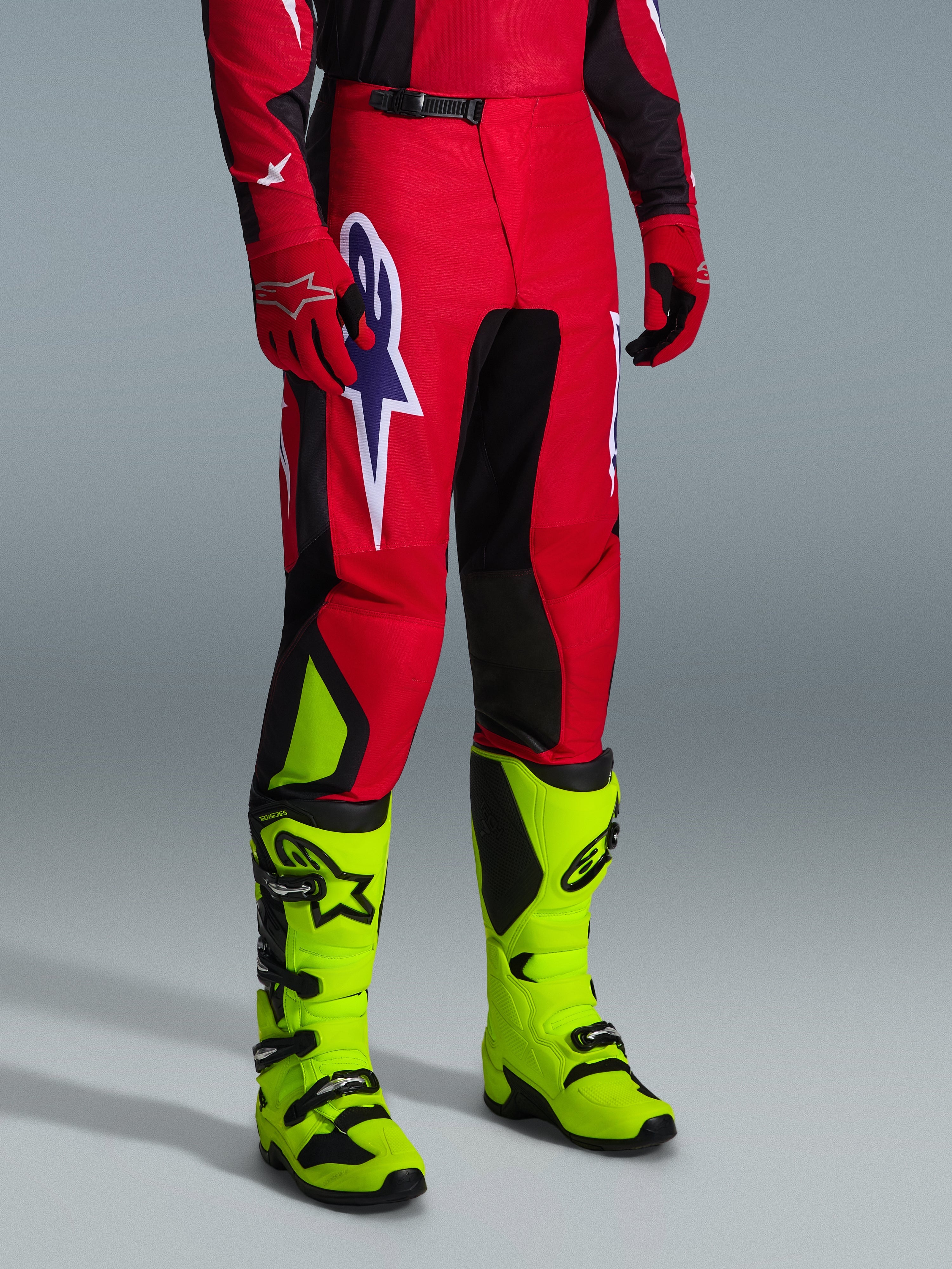 Alpinestars 2026 Fluid Grid Pants, spodnie motocrossowe, czerwone, czarne i fioletowe, noszone z neonowo żółtymi butami i czerwonymi rękawicami, z czarnymi panelami i dużym fioletowo-białym logo gwiazdy, wytrzymały sportowy krój do wyścigów terenowych