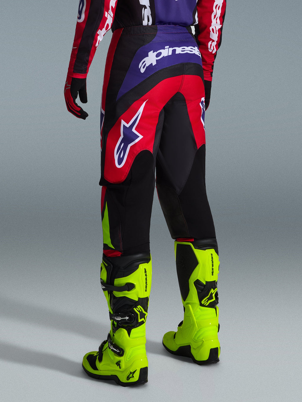 Alpinestars 2026 Fluid Grid Pants, spodnie motocrossowe, czerwone, czarne i fioletowe, widok z tyłu z dużym białym logo, zestawione z neonowożółtymi butami wyścigowymi i pasującą koszulką, wytrzymała konstrukcja o sportowym kroju zapewniająca wydajność w terenie