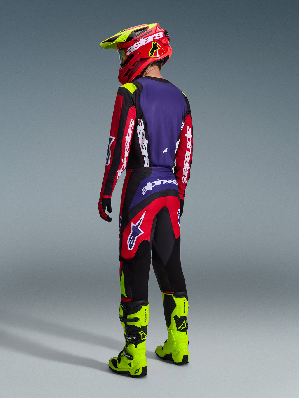 Spodnie Alpinestars Fluid Grid 2026, spodnie motocrossowe, czerwone, czarne i fioletowe, prezentowane przez motocyklistę od tyłu w pasującej koszulce i neonowożółtych butach, wyrazisty branding i ergonomiczny krój do jazdy off-road.