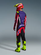 Spodnie Alpinestars Fluid Grid 2026, spodnie motocrossowe, czerwone, czarne i fioletowe, prezentowane przez motocyklistę od tyłu w pasującej koszulce i neonowożółtych butach, wyrazisty branding i ergonomiczny krój do jazdy off-road.