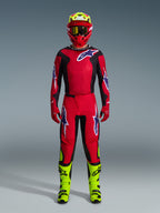 Kierowca motocrossowy w pełnym stroju wyścigowym, spodnie Alpinestars 2026 Fluid Grid w kolorze czerwonym, czarnym i fioletowym, pasująca czerwono-czarna koszulka z logo Alpinestars, czerwony kask z jaskrawożółtymi akcentami, jaskrawożółte buty i czerwone rękawice, kompletny zestaw sprzętu do wyścigów motocrossowych