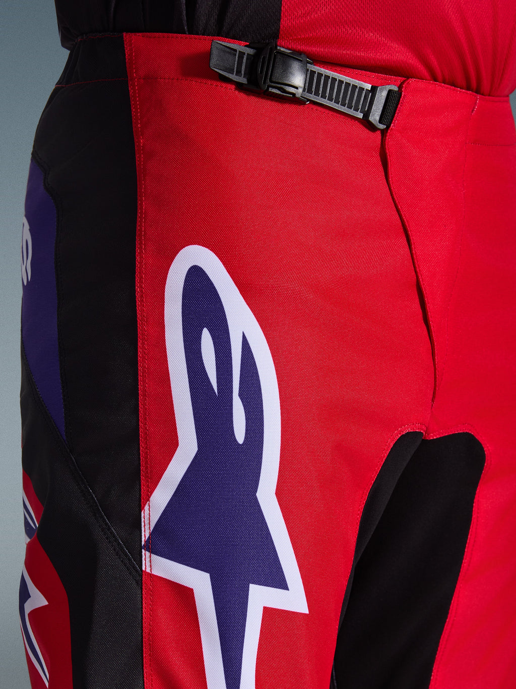 Alpinestars 2026 Fluid Grid Pants, spodnie motocrossowe, czerwone, czarne i fioletowe, zbliżenie na talię i biodra z czarnym systemem zapięcia na zapadkę i dużym fioletowym logo gwiazdy Alpinestars z białą obwódką, wytrzymała teksturowana tkanina do wyścigów terenowych.