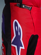 Alpinestars 2026 Fluid Grid Pants, spodnie motocrossowe, czerwone, czarne i fioletowe, zbliżenie na talię i biodra z czarnym systemem zapięcia na zapadkę i dużym fioletowym logo gwiazdy Alpinestars z białą obwódką, wytrzymała teksturowana tkanina do wyścigów terenowych.