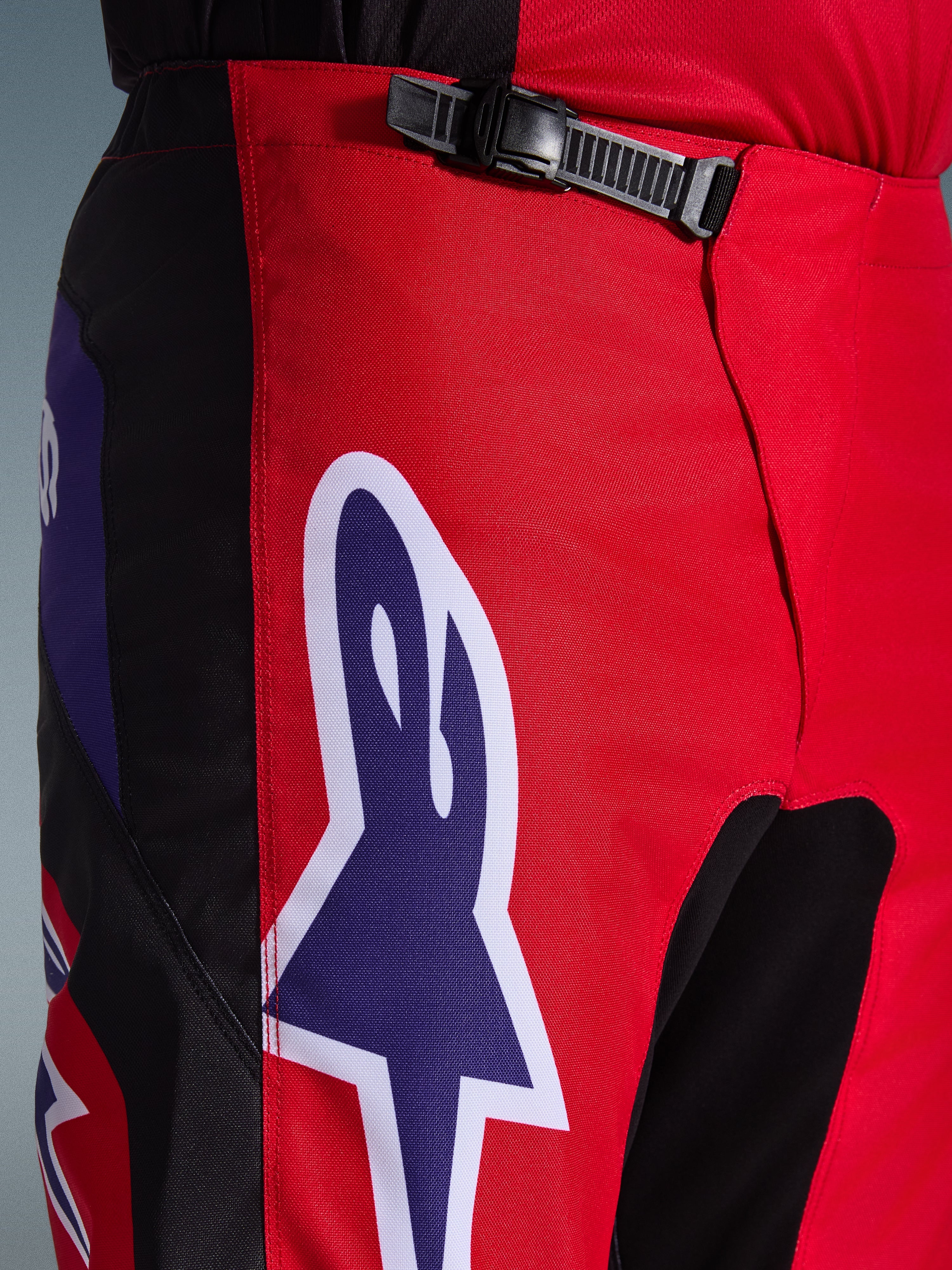 Alpinestars 2026 Fluid Grid Pants, spodnie motocrossowe, czerwone, czarne i fioletowe, zbliżenie na talię i biodra z czarnym systemem zapięcia na zapadkę i dużym fioletowym logo gwiazdy Alpinestars z białą obwódką, wytrzymała teksturowana tkanina do wyścigów terenowych.