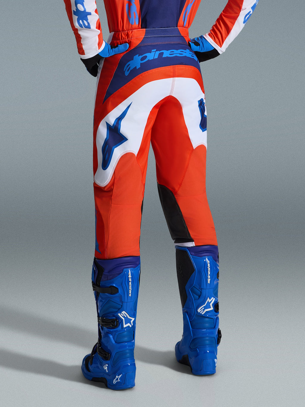 Alpinestars 2026 Fluid Grid Pants, spodnie motocrossowe, pomarańczowe i niebieskie UCLA z białymi panelami, widok z tyłu pokazujący wyraźne niebieskie logo na siedzeniu i logo gwiazdy, zestawione z pasującymi niebieskimi butami motocrossowymi, profesjonalny sprzęt do wyścigów off-road