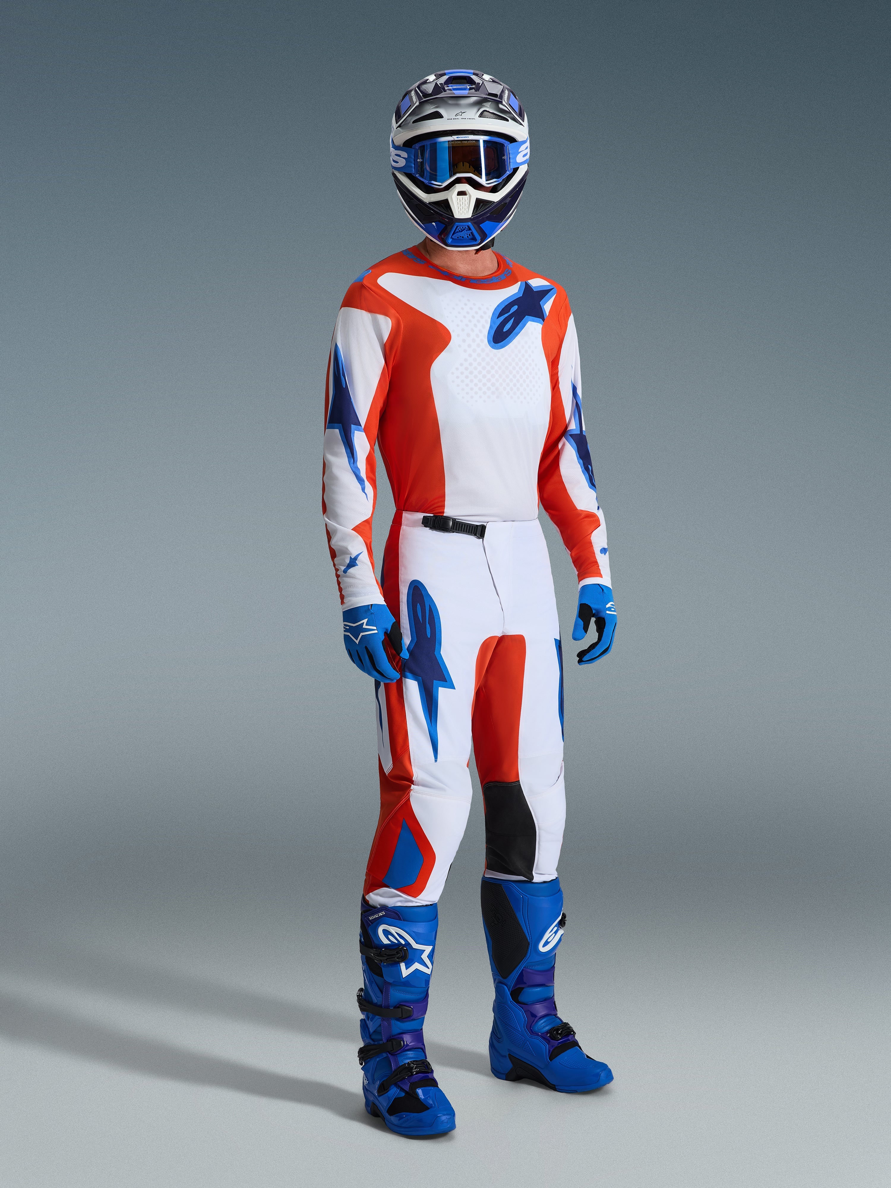 Motocyklista motocrossowy w spodniach Alpinestars 2026 Fluid Grid, pomarańczowo-niebieskie (Orange UCLA Blue), biała baza z pomarańczowymi i niebieskimi akcentami w kształcie gwiazd, pasująca koszulka z długim rękawem, niebieskie buty i kask, profesjonalny zestaw wyścigowy do jazdy off-road