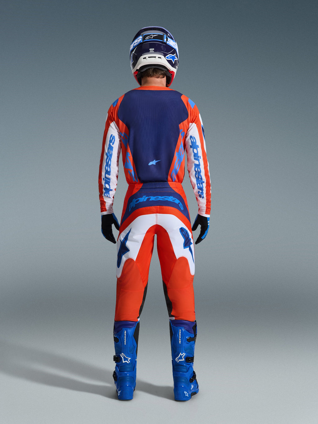 Alpinestars 2026 Fluid Grid Pants, spodnie motocrossowe, pomarańczowe i niebieskie UCLA Blue, pełny widok od tyłu na motocyklistę w kompletnym zestawie odzieży wyścigowej z pasującą koszulką, niebieskimi butami i kaskiem, z wyraźnym brandingiem i ergonomicznymi panelami do jazdy w terenie.