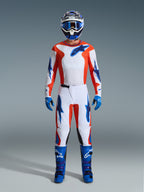 Zawodnik motocrossowy w pełnym stroju wyścigowym, spodnie Alpinestars 2026 Fluid Grid w kolorze pomarańczowym i UCLA Blue, biała koszulka z pomarańczowymi rękawami i niebieskim logo, dopasowany niebieski kask i gogle, niebieskie buty i niebieskie rękawice, skoordynowany zestaw do wyścigów off-road