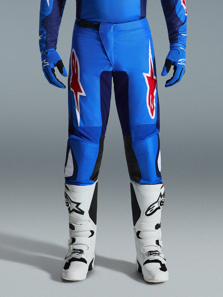 Alpinestars 2026 Fluid Grid Pants, Motocross Pants, Blue and Red, featuring bold red and white star logos on the thighs, worn with matching blue gloves and white off-road boots, athletic fit with durable panels for racing.