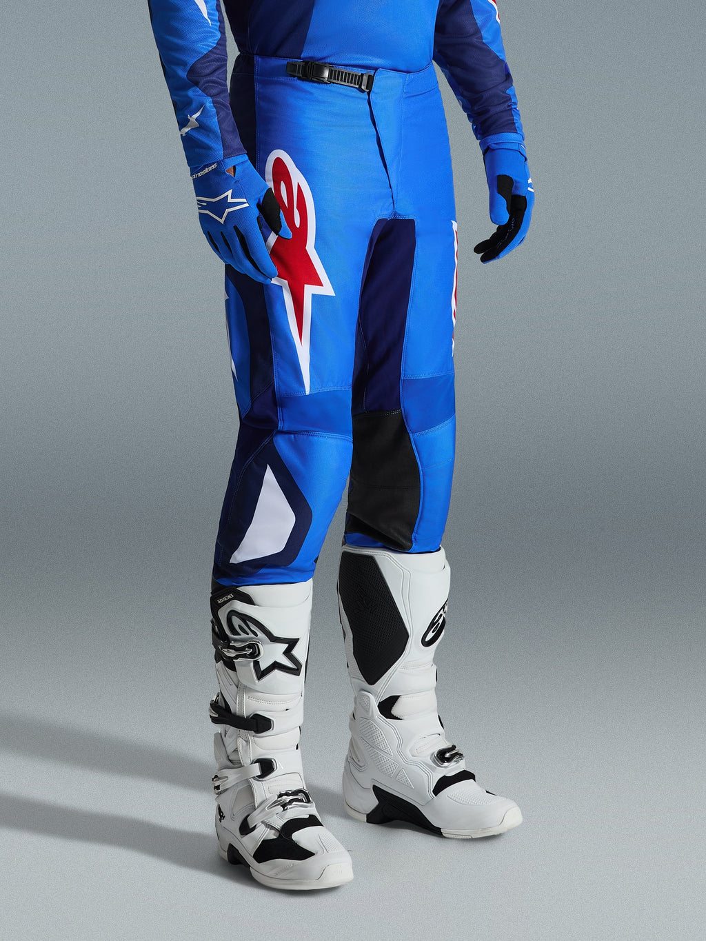 Alpinestars 2026 Fluid Grid Pants, spodnie motocrossowe, niebiesko-czerwone, zestawione z białymi butami motocrossowymi i niebieskimi rękawicami, z dużym czerwonym logo gwiazdy na udzie i czarnymi panelami po wewnętrznej stronie kolan, przeznaczone do jazdy off-road.