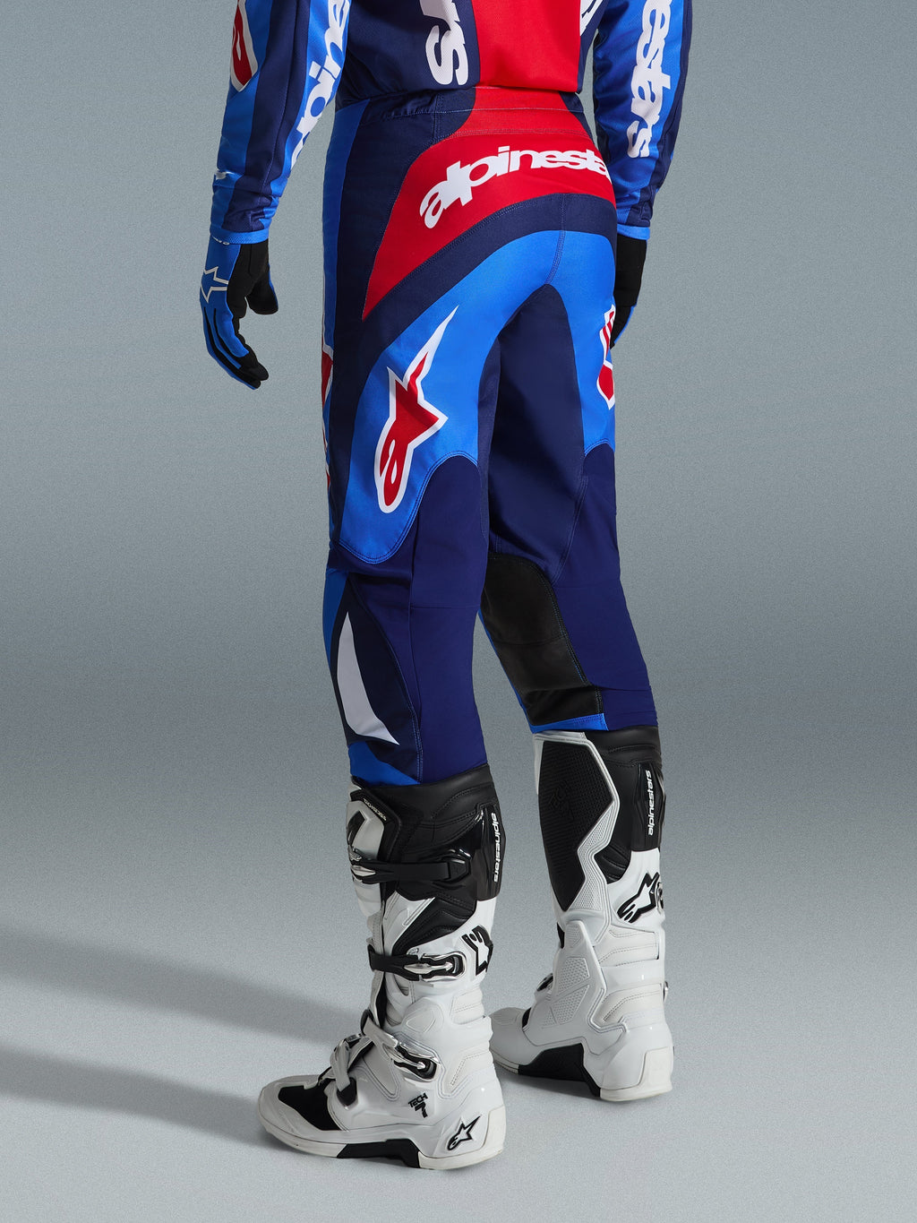 Alpinestars 2026 Fluid Grid Pants, spodnie motocrossowe, niebiesko-czerwone, widok z tyłu prezentujący wyraźne logo Alpinestars i ergonomiczną konstrukcję, noszone z biało-czarnymi butami Tech 7 i dopasowaną koszulką, wysokiej klasy odzież do jazdy terenowej