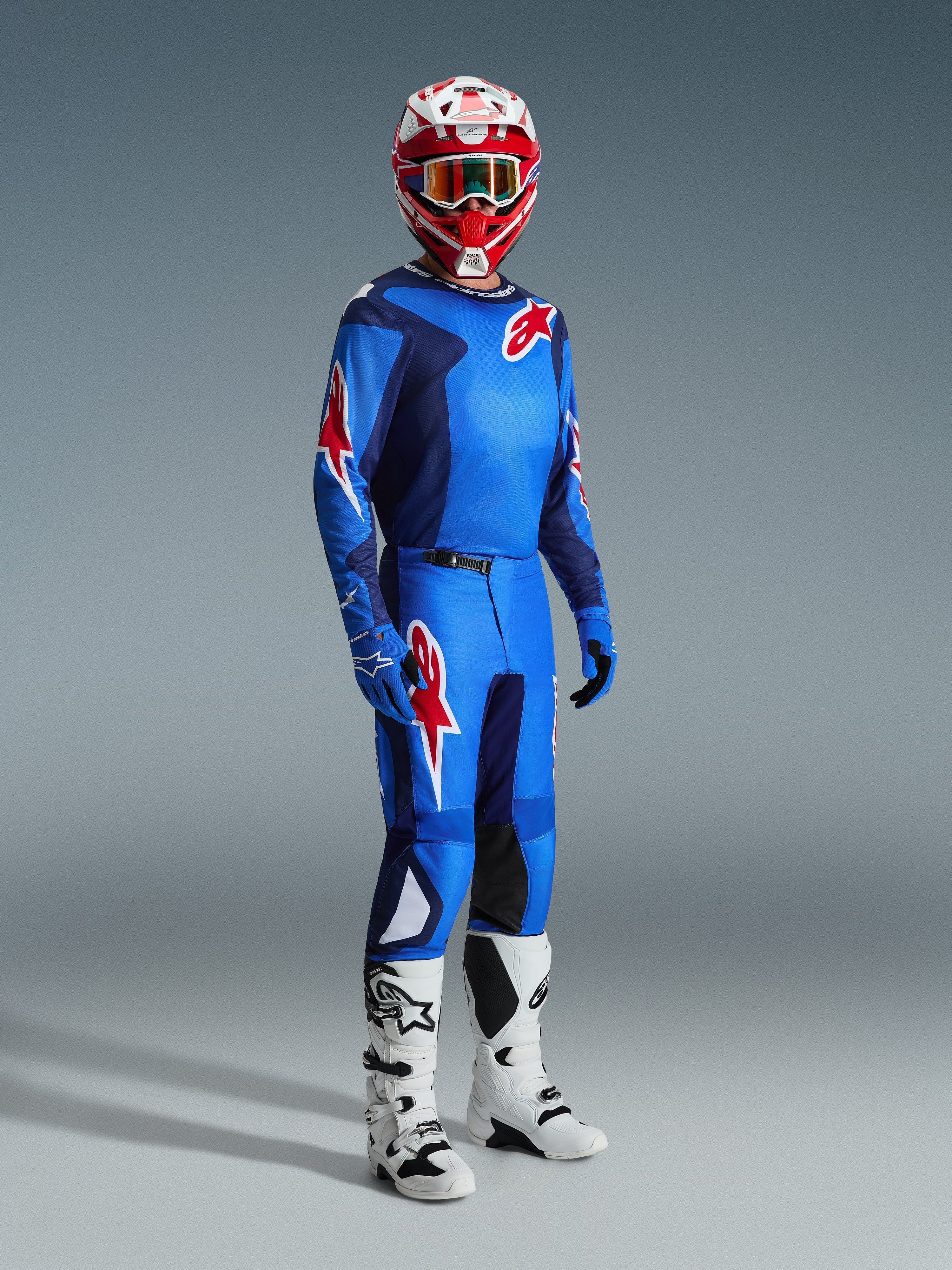 Rider motocrossowy Alpinestars w pełnym stroju wyścigowym, niebiesko-czerwona koszulka z wzorem siatki i logo, spodnie Fluid Grid 2026 w kolorze niebiesko-czerwonym z czarnymi panelami wewnętrznymi na kolanach, czerwono-biały kask z goglami, biało-czarne buty, niebieskie rękawice, kompletny zestaw do wyścigów terenowych