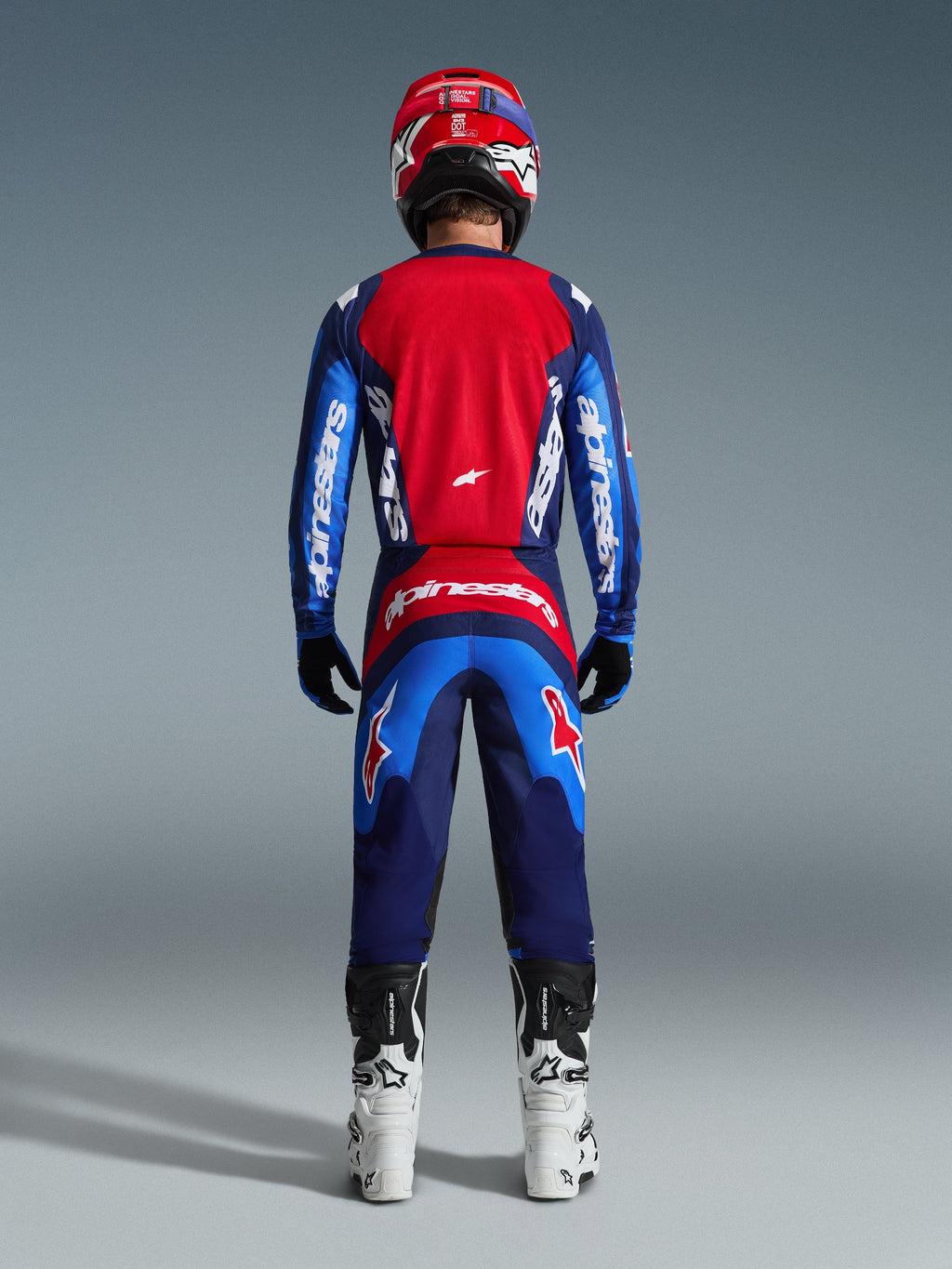 Zawodnik motocrossowy od tyłu w pełnym stroju wyścigowym, spodnie 2026 Fluid Grid w kolorze Blue Red, pasująca czerwono-niebieska koszulka z brandingiem Alpinestars, czerwono-niebieski kask, biało-czarne buty, dopasowany strój do wyścigów off-road