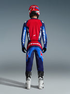 Zawodnik motocrossowy od tyłu w pełnym stroju wyścigowym, spodnie 2026 Fluid Grid w kolorze Blue Red, pasująca czerwono-niebieska koszulka z brandingiem Alpinestars, czerwono-niebieski kask, biało-czarne buty, dopasowany strój do wyścigów off-road