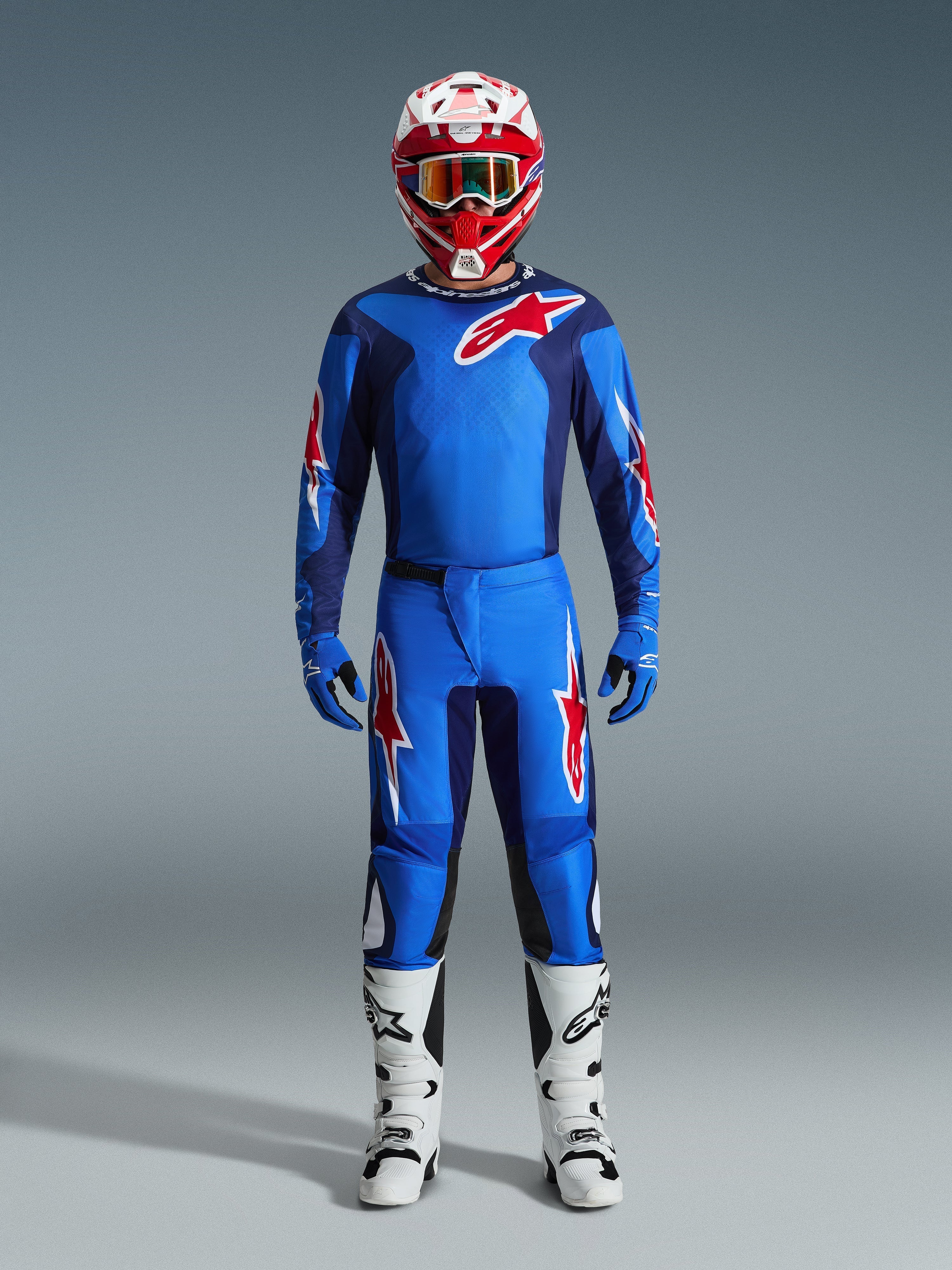 Motocyklista motocrossowy w spodniach Alpinestars 2026 Fluid Grid w kolorze niebiesko-czerwonym, dopasowana niebieska koszulka z czerwonym logo Astars, czerwono-biały kask z goglami, niebieskie rękawice i białe buty motocrossowe, kompletny zestaw wyścigowy off-road