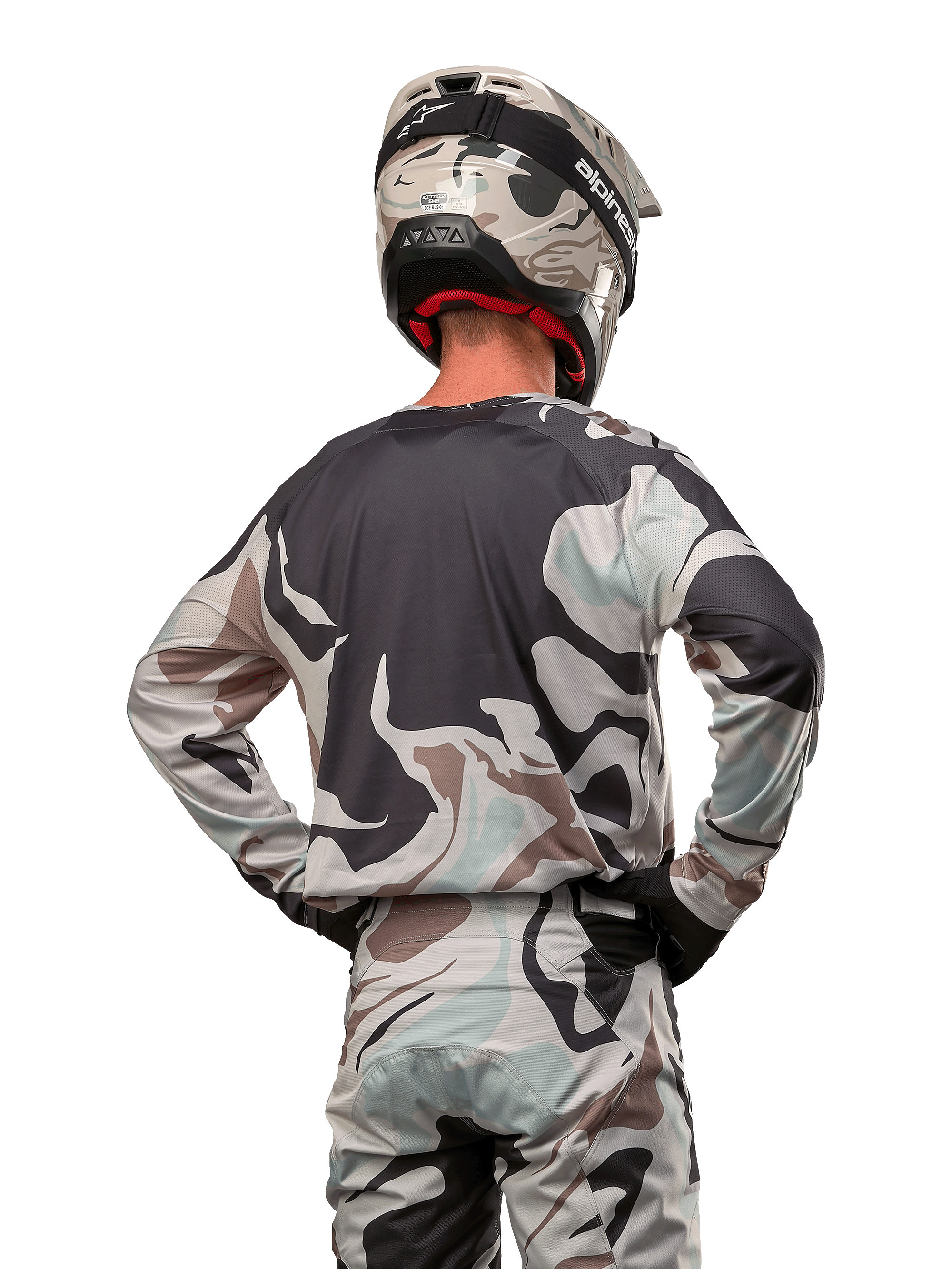 Zawodnik motocrossowy widziany od tyłu w koszulce Alpinestars 2024 Racer Tactical w kolorze Iron Camo Dust Gray, pasujące spodnie i kask z motywem kamuflażu, ręce na biodrach, dopasowany strój do wyścigów terenowych.