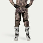 Alpinestars 2024 Racer Tactical Pants, spodnie motocrossowe, kolor Military Green Camo Brown, z dużymi białymi logotypami Alpinestars na udach i czarnymi panelami wewnątrz nogawek, noszone z biało-czarnymi butami motocrossowymi i czarnymi rękawicami, pasująca koszulka moro, widok z przodu.