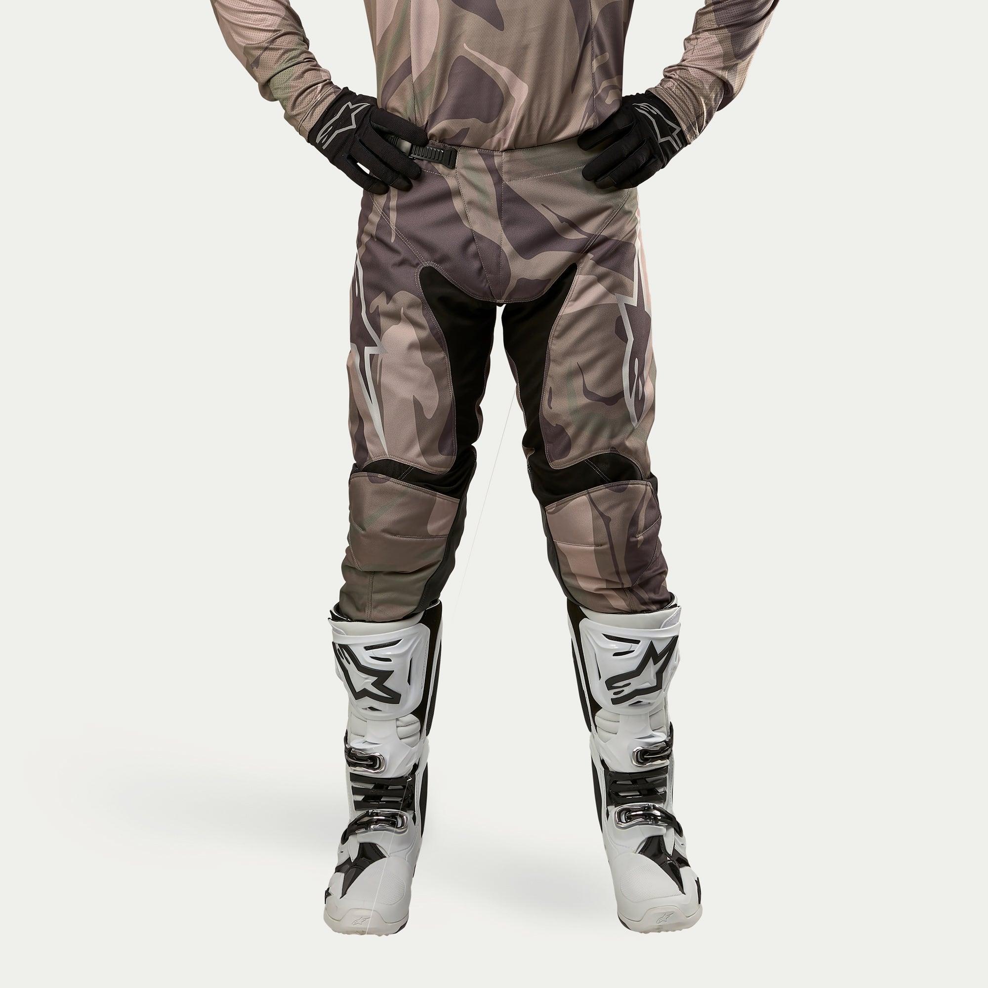 Alpinestars 2024 Racer Tactical Pants, spodnie motocrossowe, kolor Military Green Camo Brown, z dużymi białymi logotypami Alpinestars na udach i czarnymi panelami wewnątrz nogawek, noszone z biało-czarnymi butami motocrossowymi i czarnymi rękawicami, pasująca koszulka moro, widok z przodu.