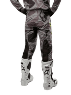 Alpinestars 2024 Racer Tactical Pants, spodnie motocrossowe, kolor Cast Gray Camo Magnet, widok z tyłu ukazujący wzór kamuflażu i czarne, oddychające strefy elastyczne, noszone z biało-czarnymi butami motocrossowymi, zaprojektowane do wyścigów terenowych.