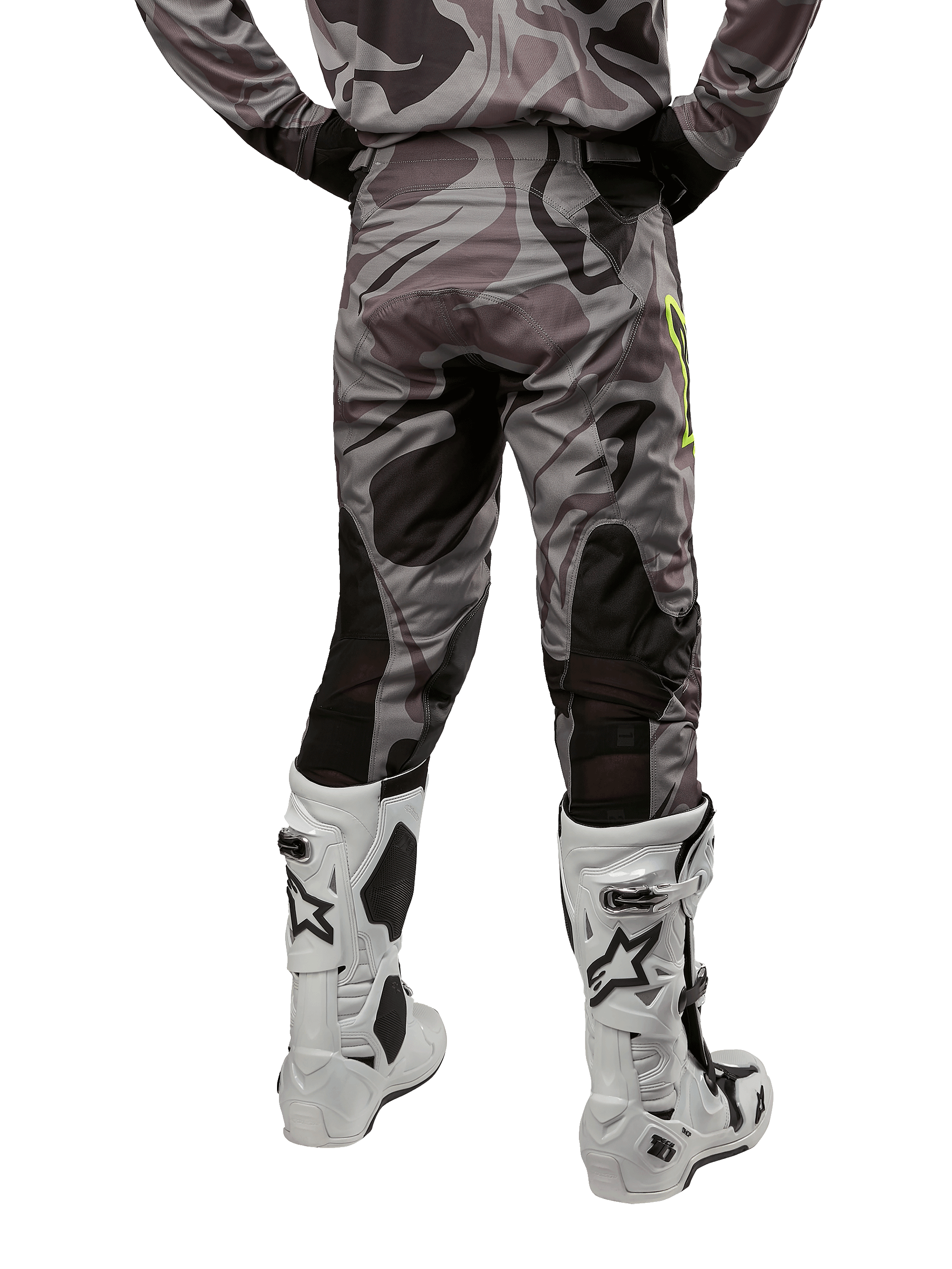 Alpinestars 2024 Racer Tactical Pants, spodnie motocrossowe, kolor Cast Gray Camo Magnet, widok z tyłu ukazujący wzór kamuflażu i czarne, oddychające strefy elastyczne, noszone z biało-czarnymi butami motocrossowymi, zaprojektowane do wyścigów terenowych.