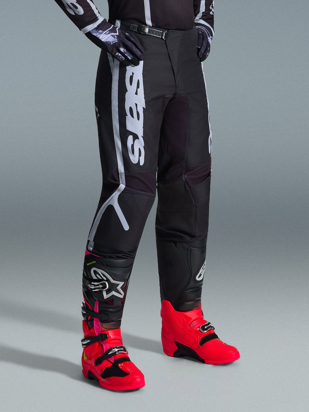 Spodnie Alpinestars Fluid Apex 2026, spodnie motocrossowe, czarno-szare, noszone z jaskraworóżowymi i czerwonymi butami oraz pasującą czarno-białą bluzą, wyraźny pionowy napis Alpinestars na udzie, wzmocnione panele i zapięcie na zapadkę w pasie, wysoka wydajność off-road