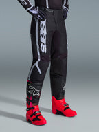 Spodnie Alpinestars Fluid Apex 2026, spodnie motocrossowe, czarno-szare, noszone z jaskraworóżowymi i czerwonymi butami oraz pasującą czarno-białą bluzą, wyraźny pionowy napis Alpinestars na udzie, wzmocnione panele i zapięcie na zapadkę w pasie, wysoka wydajność off-road