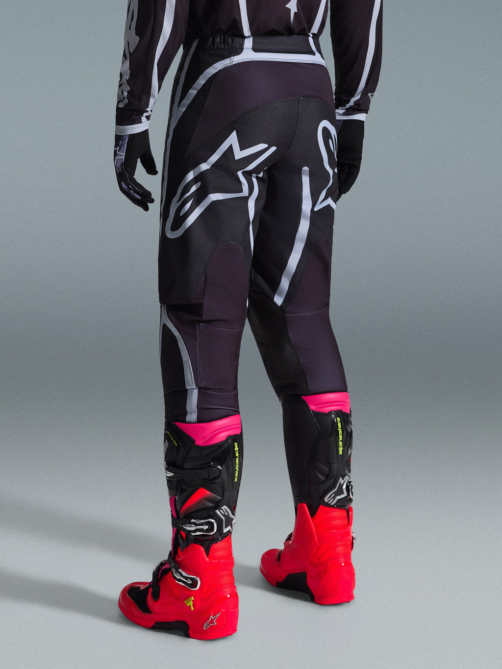 Spodnie Alpinestars 2026 Fluid Apex, spodnie motocrossowe, czarno-szare, widok z tyłu z dużymi białymi logotypami Alpinestars na siedzeniu, noszone z jaskrawoczerwonymi i różowymi butami, wytrzymała konstrukcja z materiału polyfabric 600D zapewniająca wydajność off-road