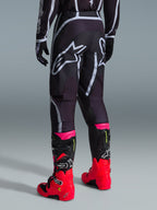 Spodnie Alpinestars 2026 Fluid Apex, spodnie motocrossowe, czarno-szare, widok z tyłu z dużymi białymi logotypami Alpinestars na siedzeniu, noszone z jaskrawoczerwonymi i różowymi butami, wytrzymała konstrukcja z materiału polyfabric 600D zapewniająca wydajność off-road