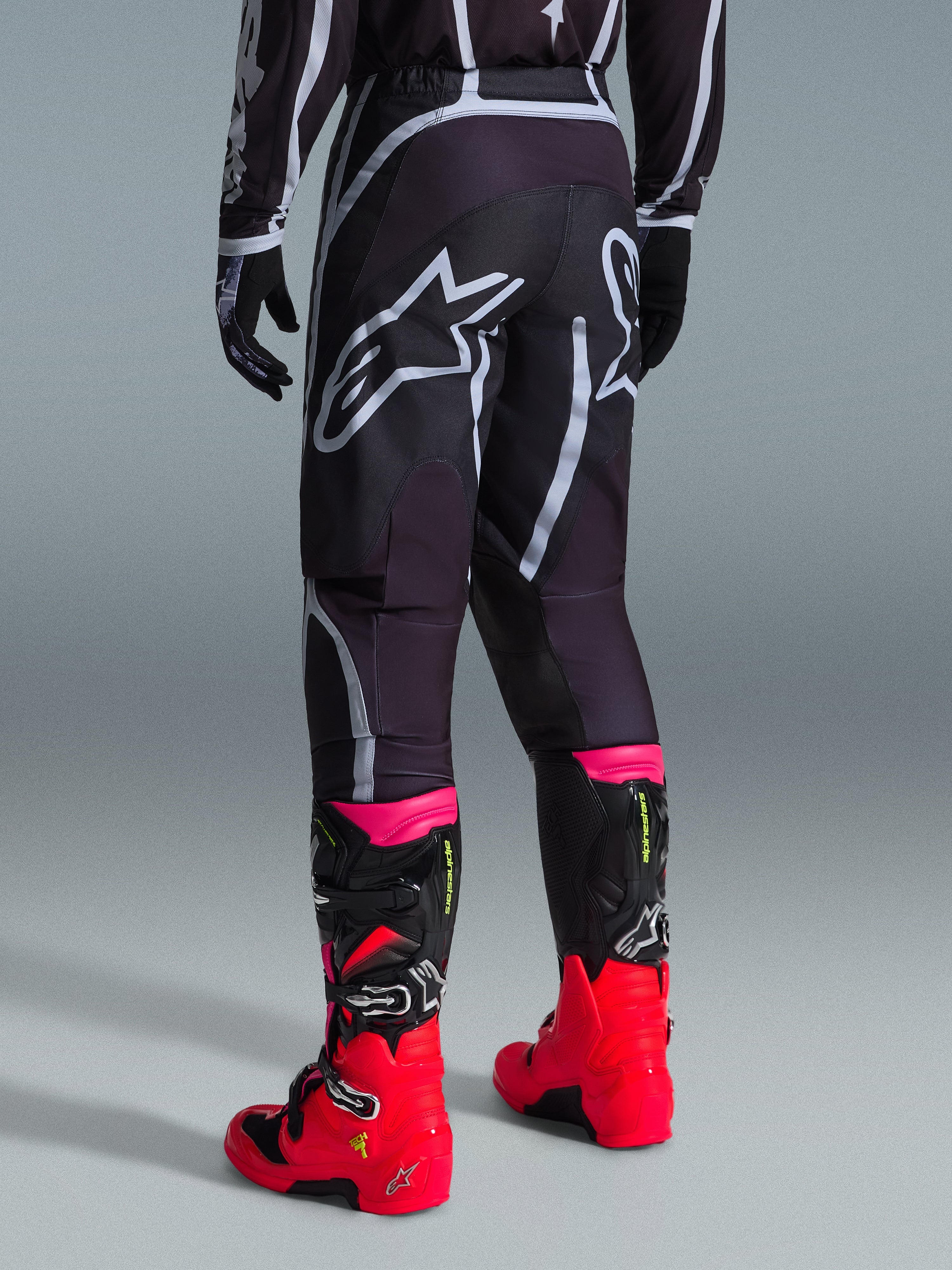 Spodnie Alpinestars 2026 Fluid Apex, spodnie motocrossowe, czarno-szare, widok z tyłu z dużymi białymi logotypami Alpinestars na siedzeniu, noszone z jaskrawoczerwonymi i różowymi butami, wytrzymała konstrukcja z materiału polyfabric 600D zapewniająca wydajność off-road