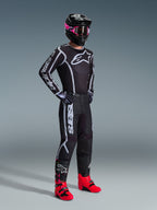 Alpinestars 2026 Fluid Apex Pants, spodnie motocrossowe, czarno-szare, zestawione z pasującą czarno-białą koszulką, czarnym kaskiem z różowymi goglami i jaskrawoczerwonymi butami, z wyraźnym białym logo na nogawkach i sportowym krojem do jazdy terenowej.
