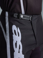 Alpinestars 2026 Fluid Apex Pants, spodnie motocrossowe, czarno-szare, zbliżenie na zintegrowany system zapięcia zapadkowego zapewniający pewne dopasowanie, biała grafika logo na wytrzymałym poliestrze 600D.