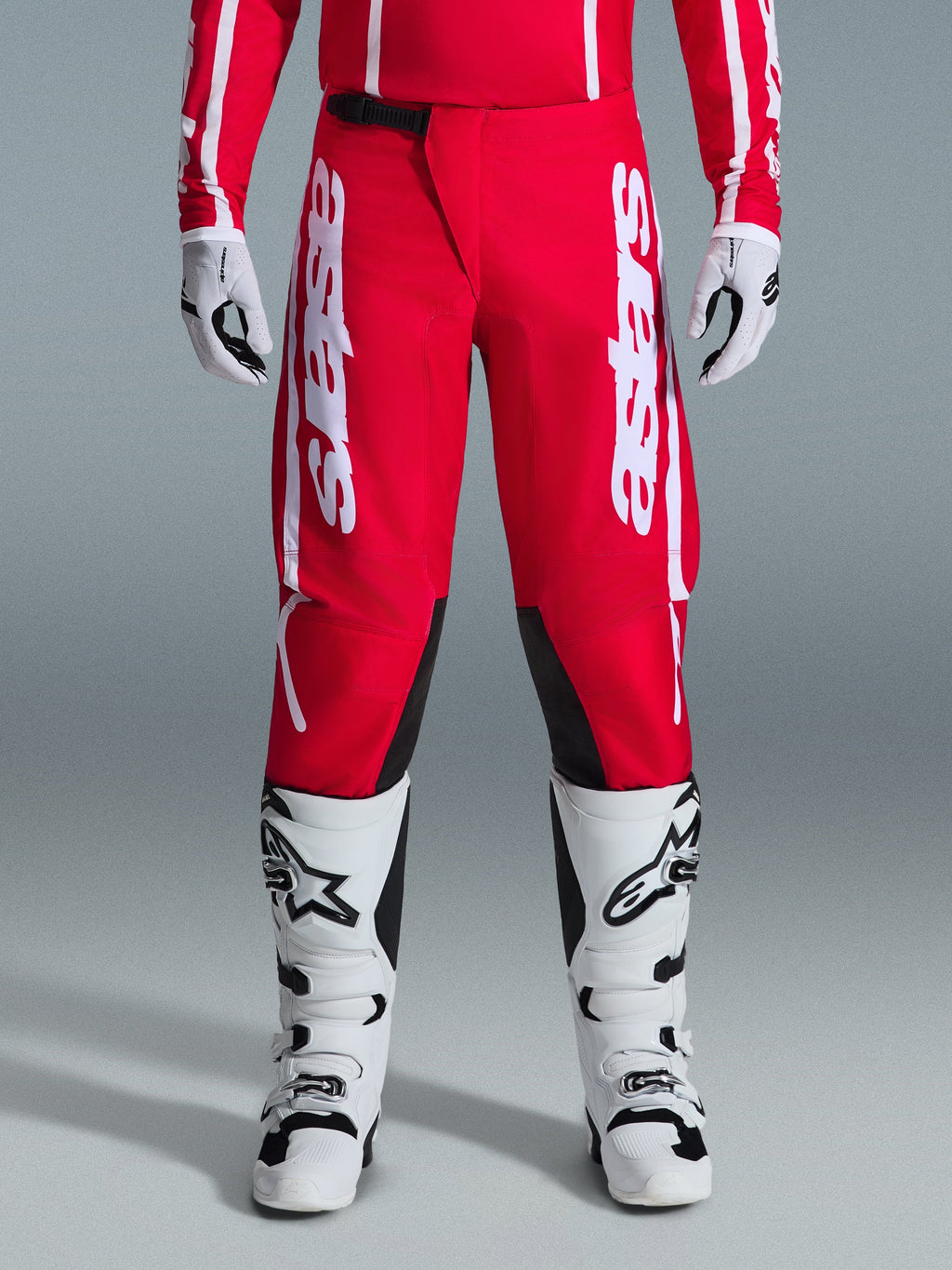 Alpinestars 2026 Fluid Apex Pants, spodnie motocrossowe, czerwono-białe, z dużymi białymi logo Astars na udach i czarnymi panelami po wewnętrznej stronie kolan, noszone z pasującymi biało-czarnymi butami i rękawicami dla pełnego zestawu wyścigowego off-road